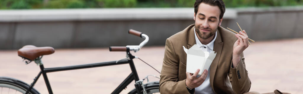 glücklicher Geschäftsmann im Anzug mit Pappschachtel mit thailändischem Essen und Essstäbchen, während er neben dem Fahrrad sitzt, Banner - Foto, Bild