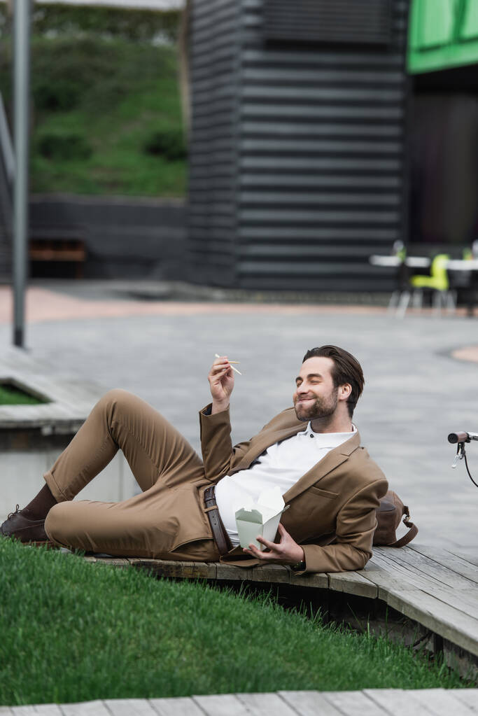 homem de negócios feliz segurando caixa de papelão com comida asiática e pauzinhos enquanto deitado perto de grama - Foto, Imagem