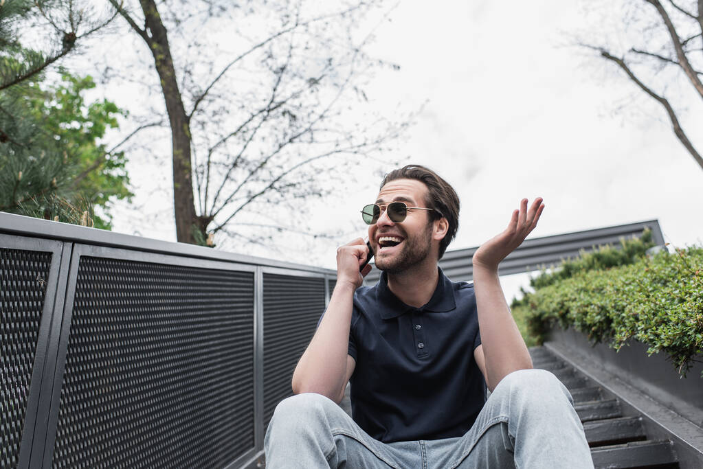 hymyilevä mies aurinkolaseissa ja poolopaidassa puhuu kännykällä ulkona  - Valokuva, kuva