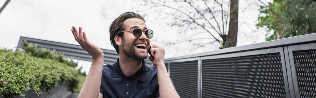 verbaasd man in zonnebril en polo shirt praten op mobiele telefoon buiten, banner  - Foto, afbeelding