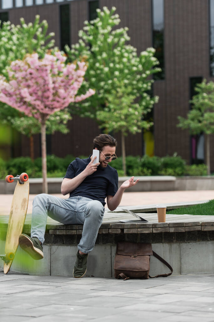 веселий чоловік в сонцезахисних окулярах розмовляє по смартфонах біля дошки і гаджетів
  - Фото, зображення