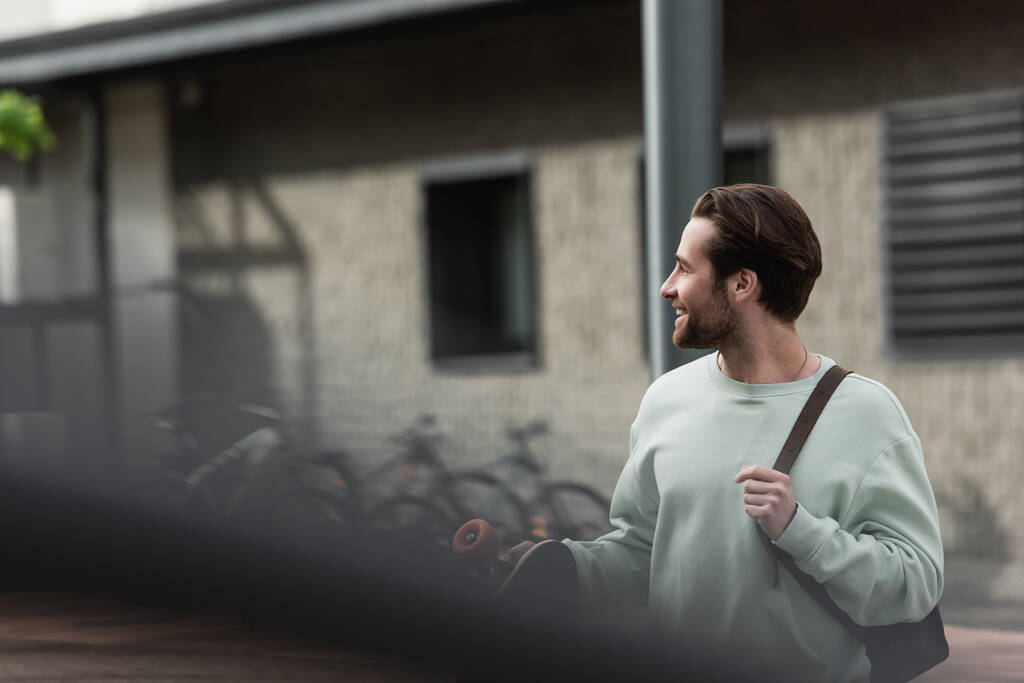 szczęśliwy brodaty mężczyzna w bluzie trzyma longboard i skórzany pasek plecaka na zewnątrz  - Zdjęcie, obraz
