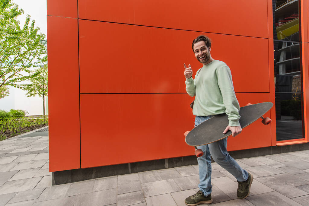 šťastný muž v mikině drží longboard a ukazuje prstem při pohledu na kameru u oranžové stěny - Fotografie, Obrázek