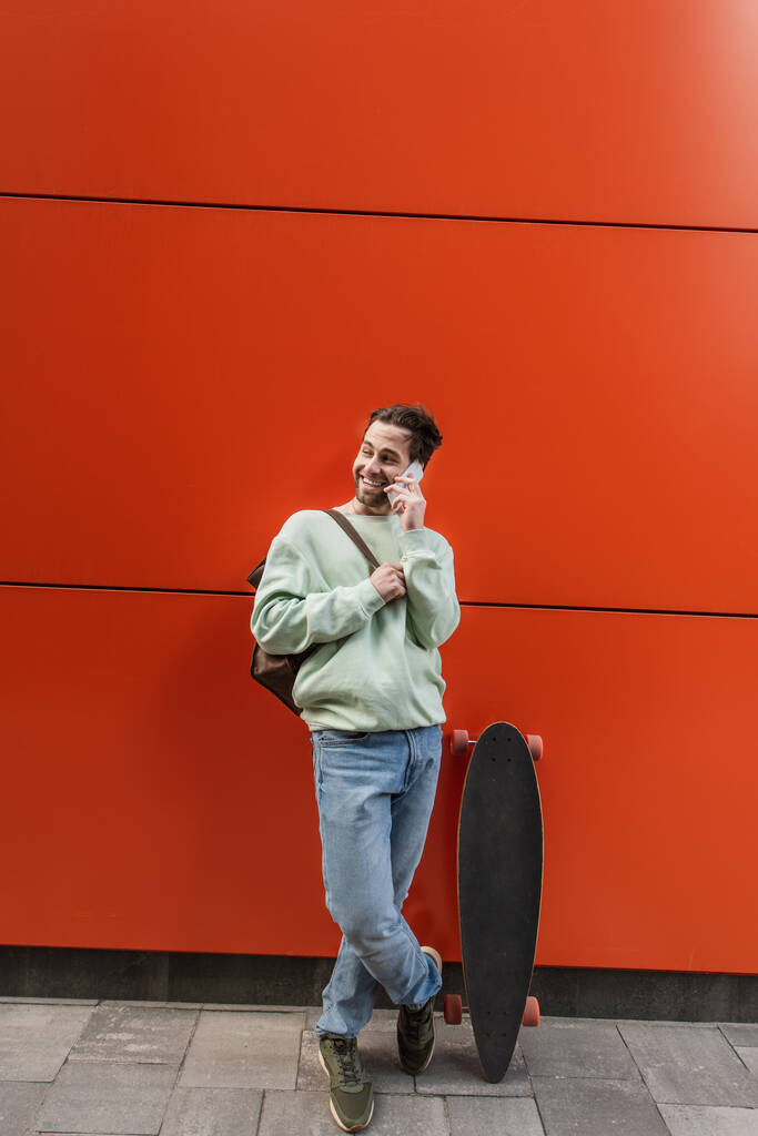 glücklicher Mann im Sweatshirt, der in der Nähe orangefarbener Mauer mit dem Handy telefoniert - Foto, Bild