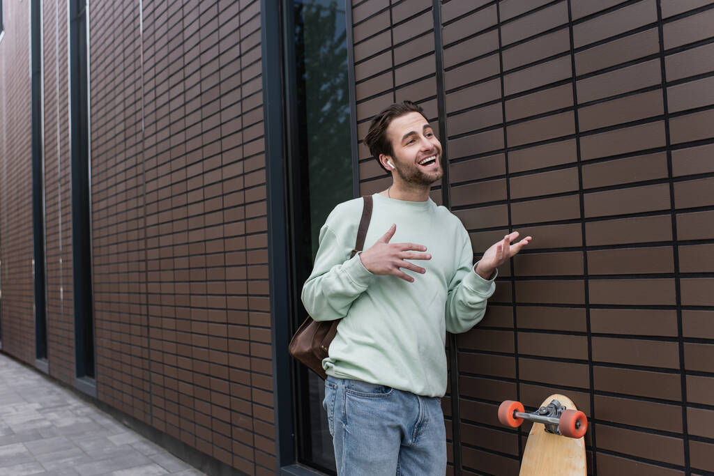 hombre alegre en sudadera y auriculares inalámbricos hablando y haciendo gestos fuera  - Foto, Imagen