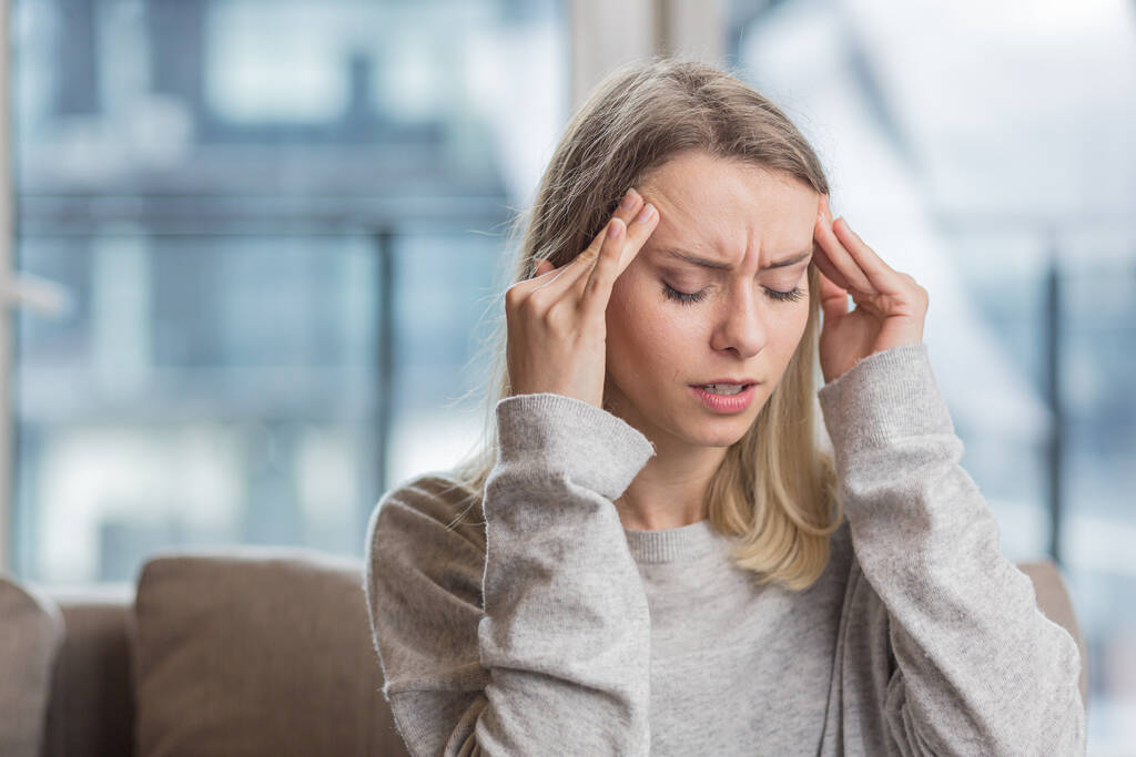Eine Frau, die ihren Kopf ängstlich über unerträgliche Kopfschmerzen hält, sitzt zu Hause auf der Couch - Foto, Bild