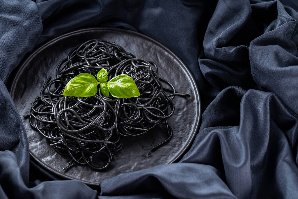 Pastas negras y albahaca sobre un fondo oscuro. Espaguetis negros con tinta de sepia sobre un paño negro. Pasta con albahaca fresca en un plato negro. Copiar espacio. Vista superior - Foto, Imagen