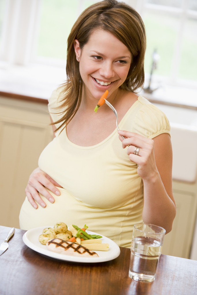 Raskaana oleva nainen keittiössä syö kanaa ja vihanneksia hymyillen
 - Valokuva, kuva