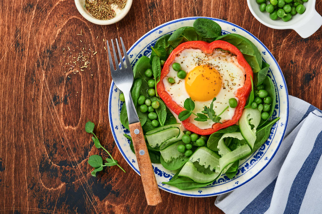 Красный перец фаршированный яйцами, листья шпината, зеленый горох и микрозелень на тарелке для завтрака на старом деревянном столе. Вид сверху. - Фото, изображение
