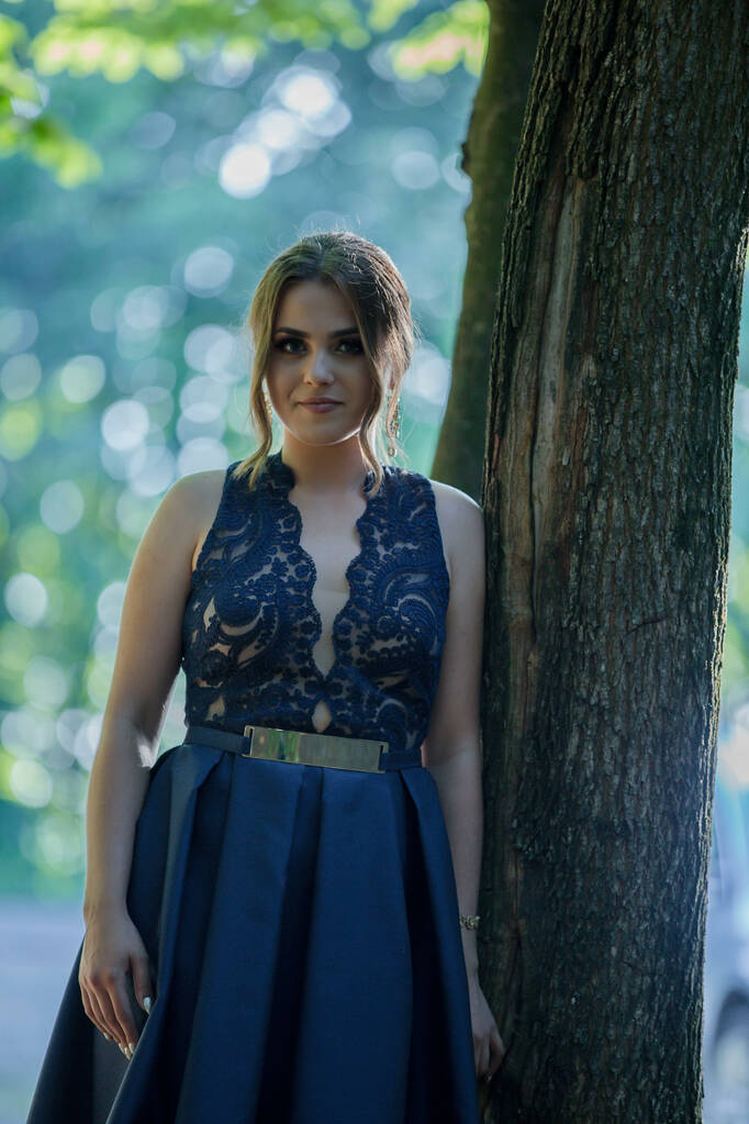Привлекательная белая женщина в элегантном голубом платье, прислонившаяся к дереву в парке - Фото, изображение