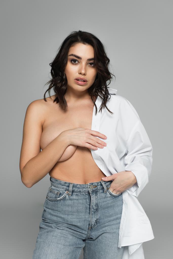 Sexy woman with naked breast holding hand in pocket of jeans isolated on grey  - Photo, Image