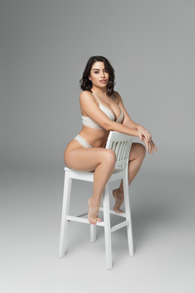 Великолепная женщина в нижнем белье позирует на белом стуле на сером фоне  - Фото, изображение