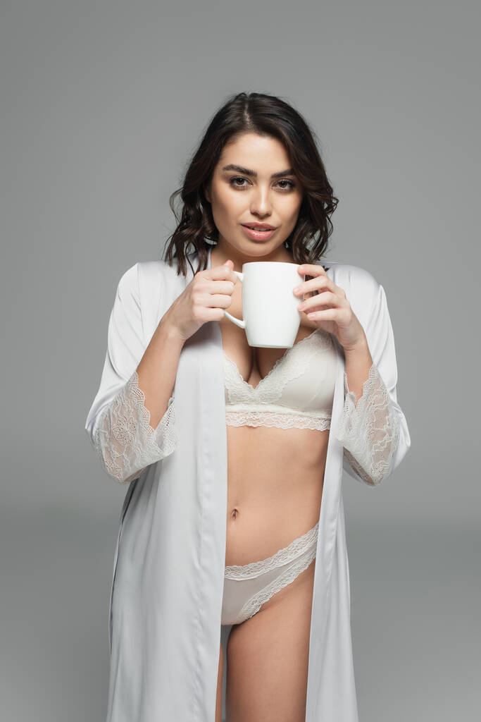 Smiling woman in silk robe and lingerie holding cup isolated on grey - Photo, Image