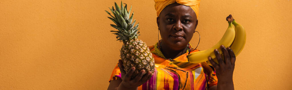 afrikkalainen amerikkalainen nainen keltainen turbaani ja värikäs mekko tilalla ananas ja banaanit oranssi, banneri - Valokuva, kuva