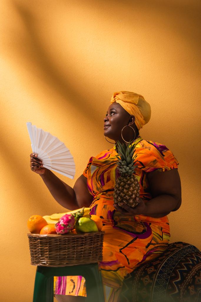 Afrikanische Amerikanerin mittleren Alters sitzt neben Früchten und hält Ananas und Fächer auf Orange - Foto, Bild