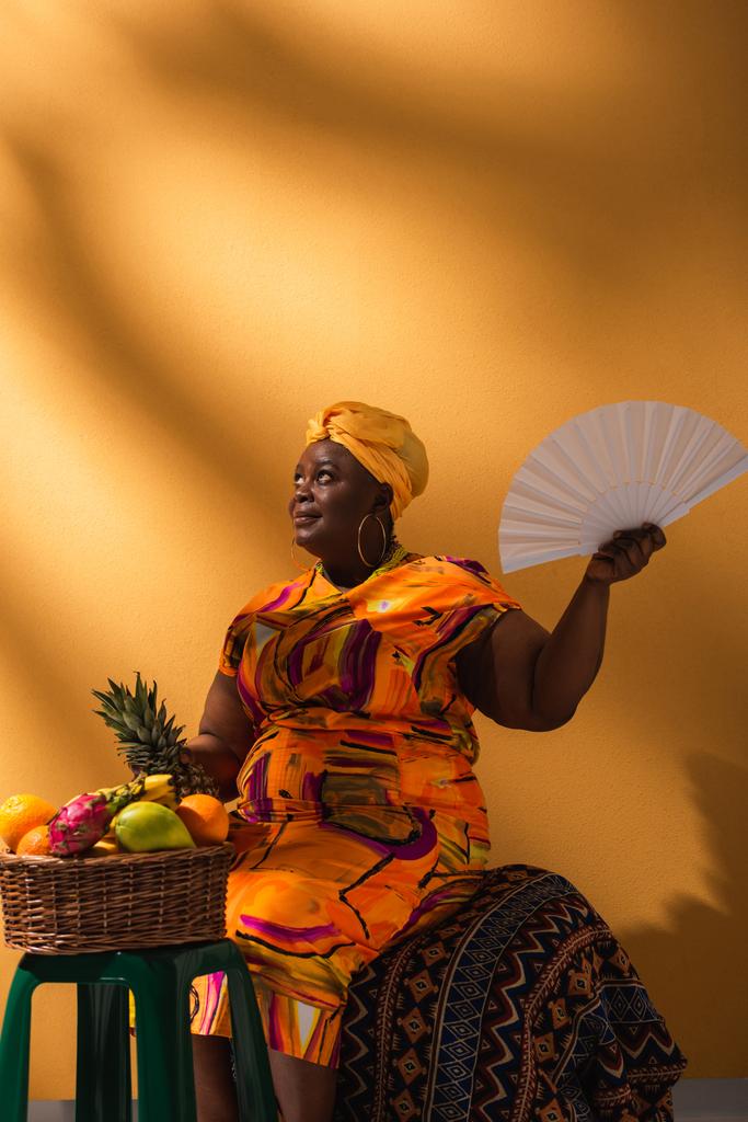 χαμογελαστή μεσήλικη Αφροαμερικανή που κάθεται κοντά σε φρούτα και κρατάει ανανά και ανεμιστήρα στο πορτοκάλι - Φωτογραφία, εικόνα