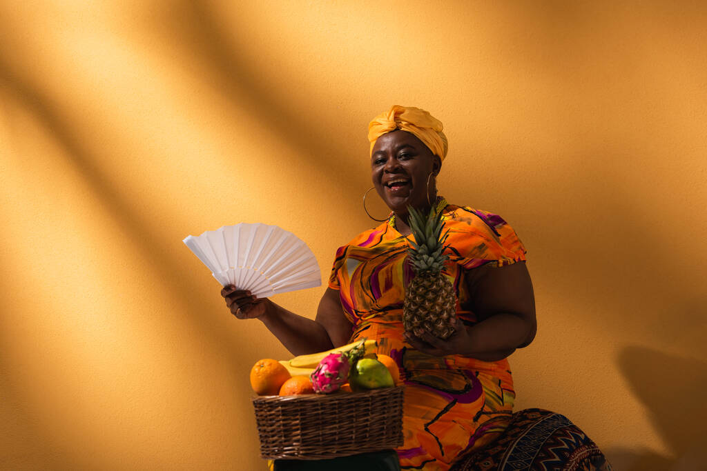 pozitivní středního věku africká americká žena sedí v blízkosti ovoce a drží ananas a ventilátor na oranžové - Fotografie, Obrázek