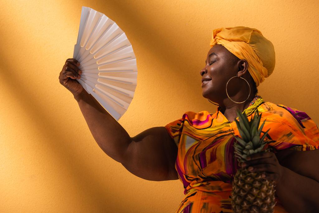 Afrikanische Amerikanerin mittleren Alters sitzt mit geschlossenen Augen, hält Ananas in der Hand und winkt mit Fächer auf Orange  - Foto, Bild