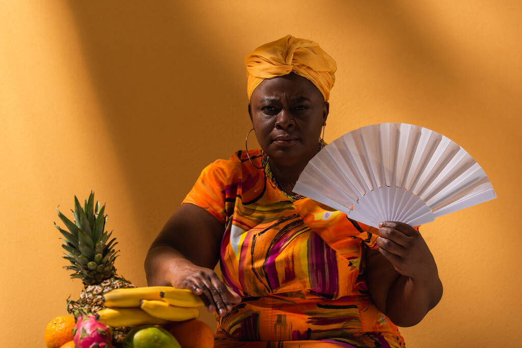 seria de mediana edad afroamericana mujer sentada con ventilador cerca de frutas en naranja - Foto, Imagen