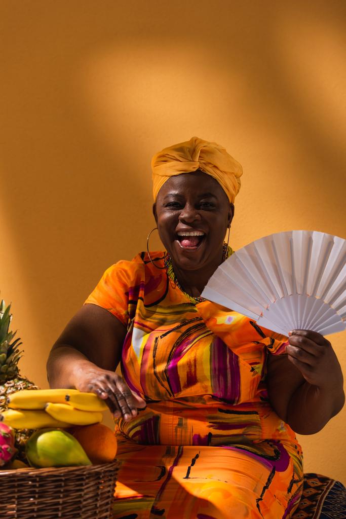 smiling middle aged african american woman sitting with fan near fruits on orange - Photo, Image