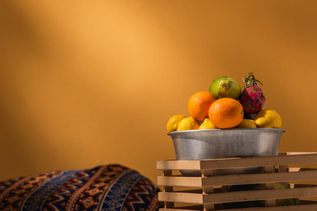 frutas exóticas en cuenco de metal y caja de madera cerca de manta de adorno en naranja - Foto, imagen