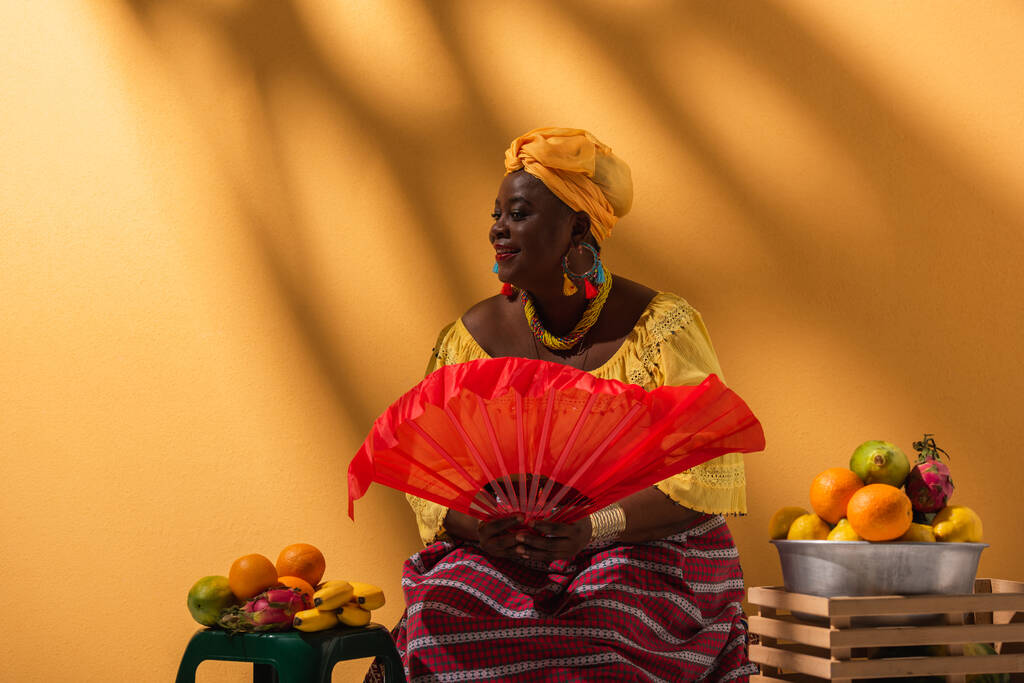 cheerful middle aged african american woman selling fruits and holding fan on orange - Photo, Image