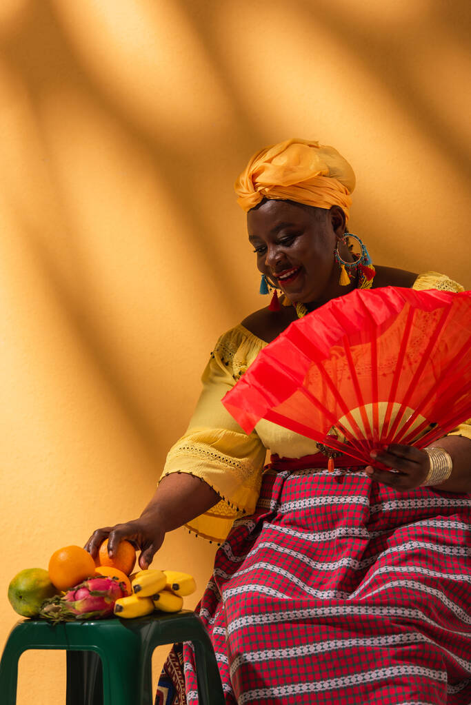 joyeuse femme afro-américaine d'âge moyen touchant des fruits et tenant ventilateur sur jaune - Photo, image