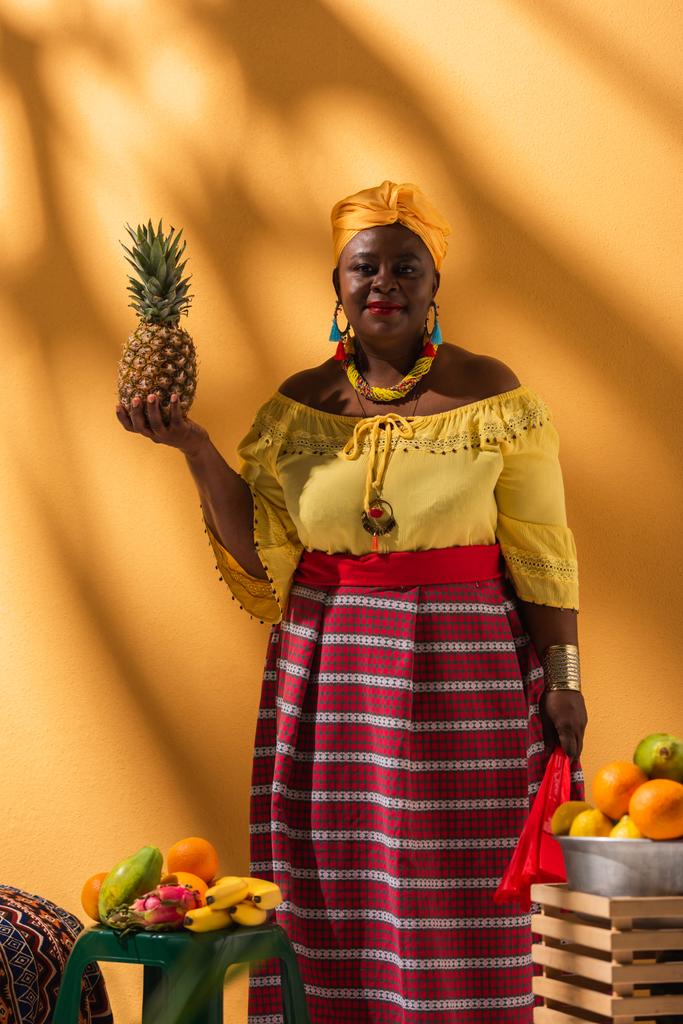 позитивна середнього віку афроамериканка, що продає фрукти з вентилятором і ананасом в руках на апельсині
 - Фото, зображення