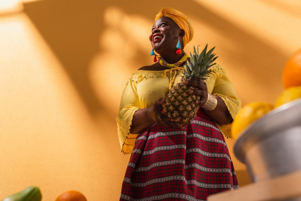 Άποψη χαμηλής γωνίας χαμογελαστή μεσήλικας Αφροαμερικανή γυναίκα που πουλάει φρούτα με ανανά στο χέρι σε πορτοκαλί - Φωτογραφία, εικόνα