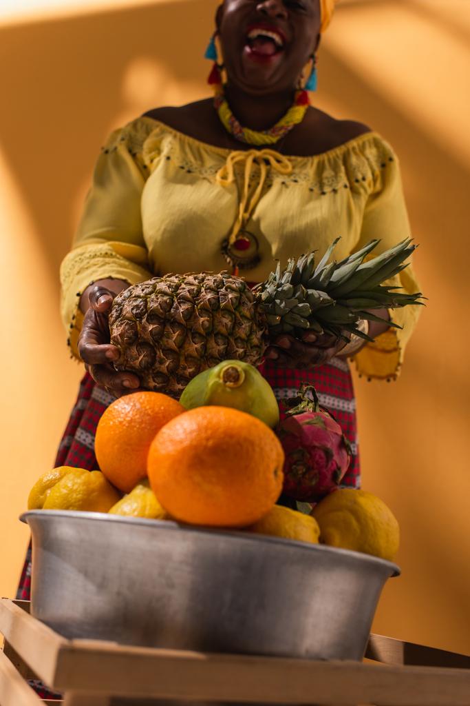 lage hoek uitzicht van vrolijke middelbare leeftijd Afrikaanse Amerikaanse vrouw verkopen fruit met ananas in handen op oranje - Foto, afbeelding