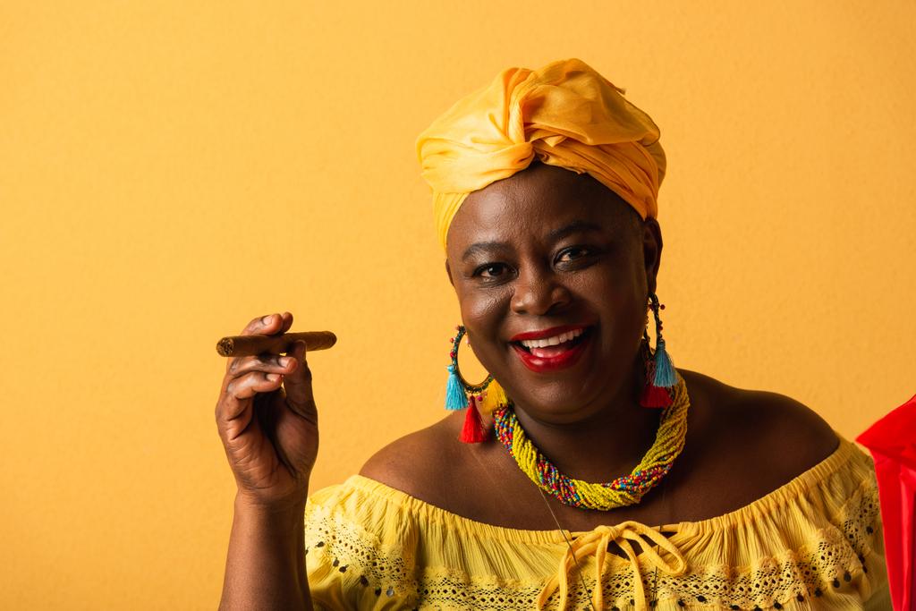 uśmiechnięta Afroamerykanka w średnim wieku w żółtym turbanie i bluzce trzymająca cygaro na żółtym - Zdjęcie, obraz