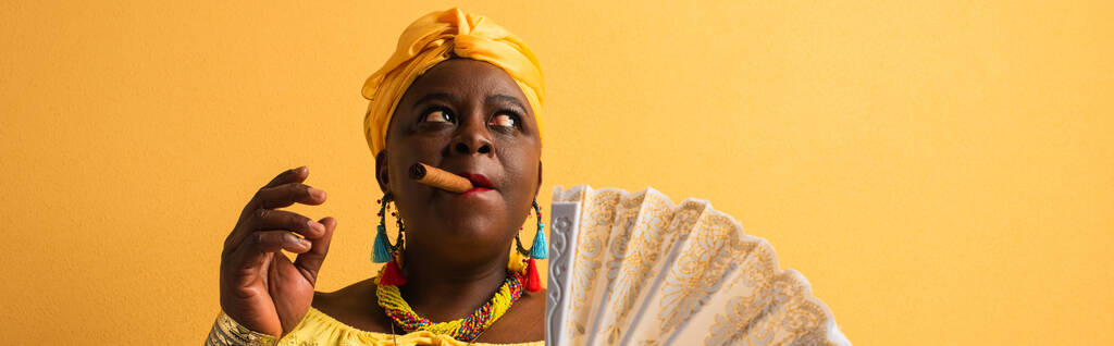 femme afro-américaine d'âge moyen en turban jaune et chemisier tenant ventilateur et cigare fumant sur jaune, bannière - Photo, image