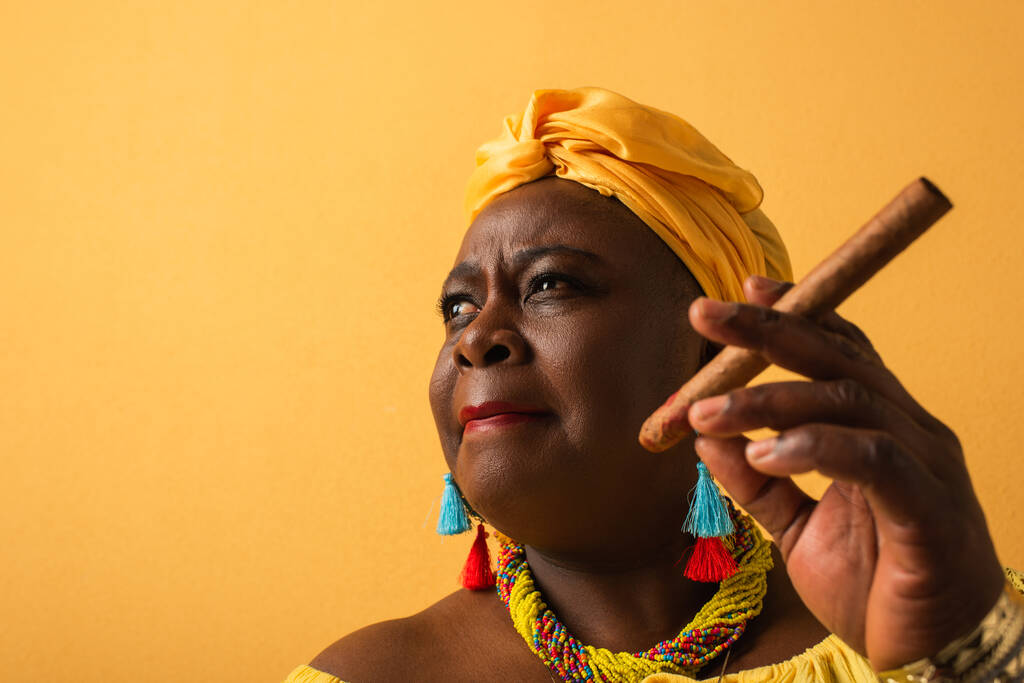 portrait d'une femme afro-américaine d'âge moyen en turban jaune tenant un cigare sur du jaune - Photo, image