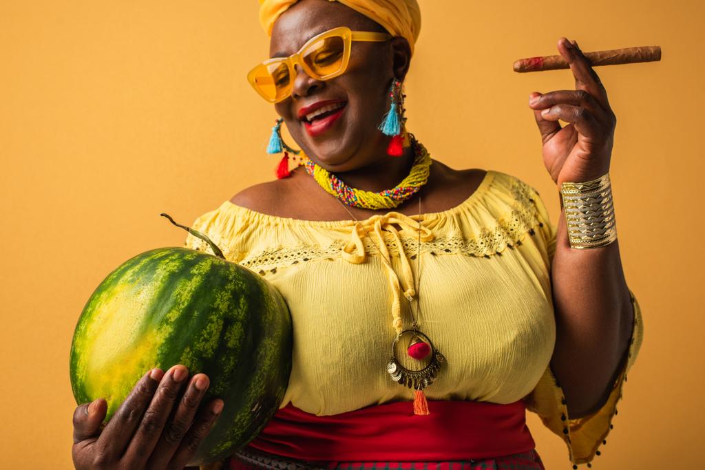 Африканская женщина средних лет в яркой одежде и солнцезащитных очках держит арбуз и сигару на желтом - Фото, изображение