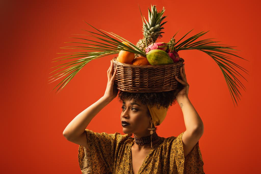mladá africká americká žena drží koš s exotickým ovocem na hlavě a dívá se jinam na červené - Fotografie, Obrázek
