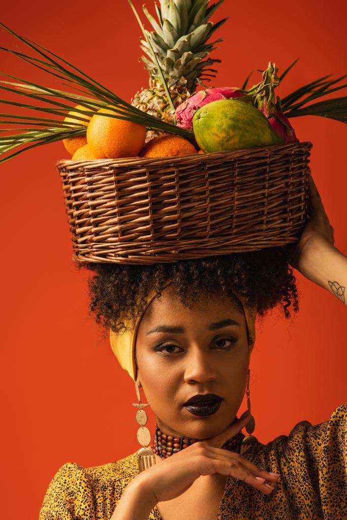 ritratto di una giovane donna afroamericana seria che tiene un cesto con frutti esotici sulla testa su rosso - Foto, immagini
