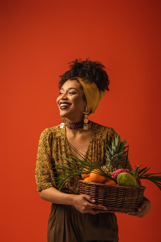 Lächelnde junge afrikanisch-amerikanische Frau hält Korb mit exotischen Früchten auf rot - Foto, Bild