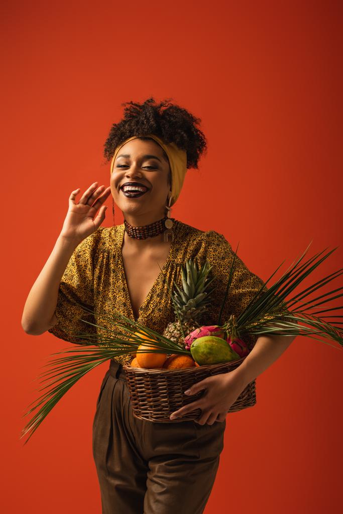 positivo joven afroamericana mujer con mano cerca de cara celebración cesta con frutas exóticas en rojo - Foto, Imagen