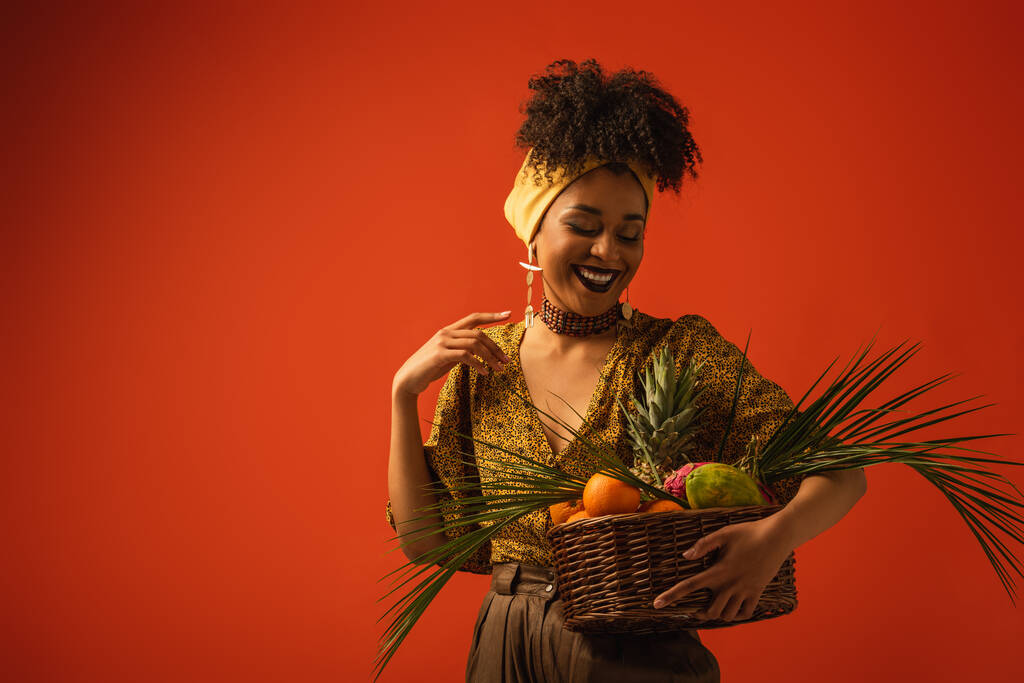 χαμογελαστή νεαρή Αφροαμερικανή γυναίκα με το χέρι κοντά στον ώμο κρατώντας καλάθι με εξωτικά φρούτα στο κόκκινο - Φωτογραφία, εικόνα