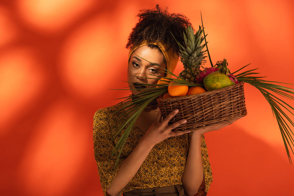 若いアフリカ系アメリカ人の女性がオレンジの頭の近くにエキゾチックな果物のバスケットを持っています - 写真・画像