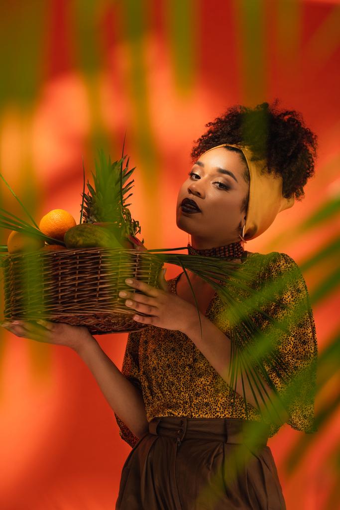 young african american woman holding basket with exotic fruits behind blurred palm leaves on orange - Photo, Image