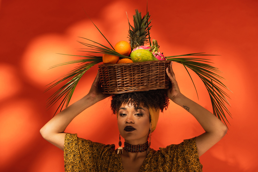 νεαρή ενήλικη Αφροαμερικανή γυναίκα που κρατά καλάθι με εξωτικά φρούτα στο κεφάλι σε πορτοκαλί - Φωτογραφία, εικόνα