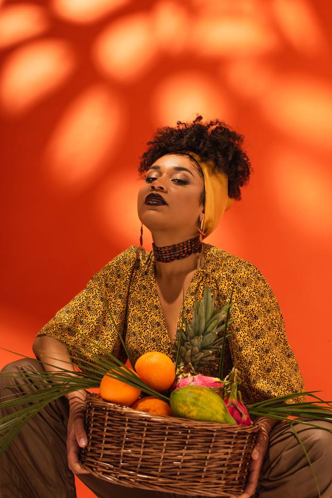 confiado joven afroamericana mujer celebración cesta con frutas exóticas en naranja - Foto, Imagen