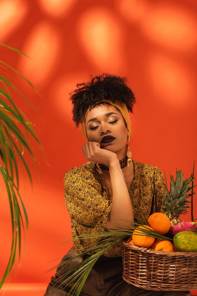young african american woman with closed eyes and hand near face sitting near basket with exotic fruits on orange - Photo, Image