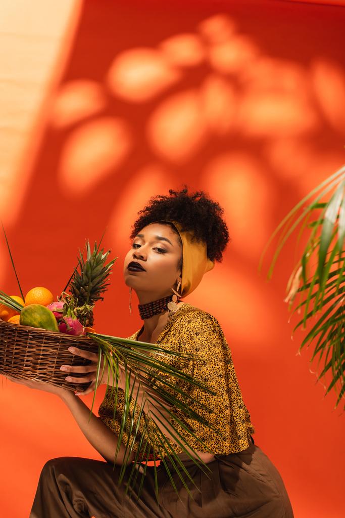 jeune femme afro-américaine tenant panier avec des fruits exotiques et regardant la caméra sur orange - Photo, image