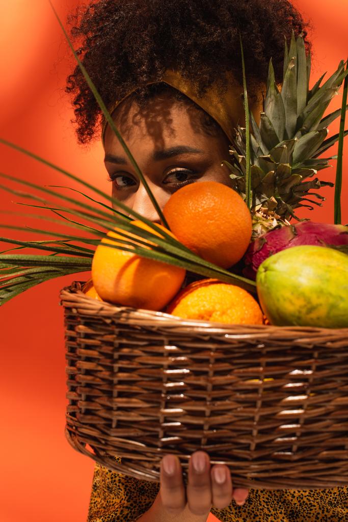 νεαρή Αφροαμερικανή γυναίκα που καλύπτει το πρόσωπο με εξωτικά φρούτα στο καλάθι με πορτοκαλί - Φωτογραφία, εικόνα