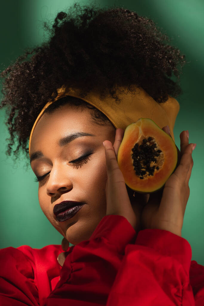 portrait de jeune femme afro-américaine avec les yeux fermés tenant la moitié de la coupe de papaye dans les mains sur vert - Photo, image
