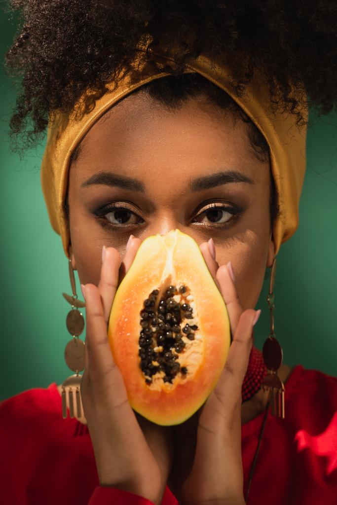 молодая африканская американка, покрывающая лицо половинкой спелой папайи на зеленом - Фото, изображение