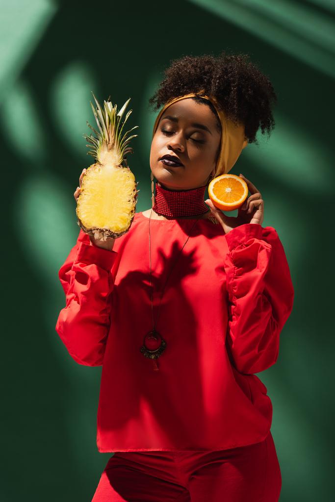 young african american woman with closed eyes holding half cut of pineapple and orange on green - Photo, Image