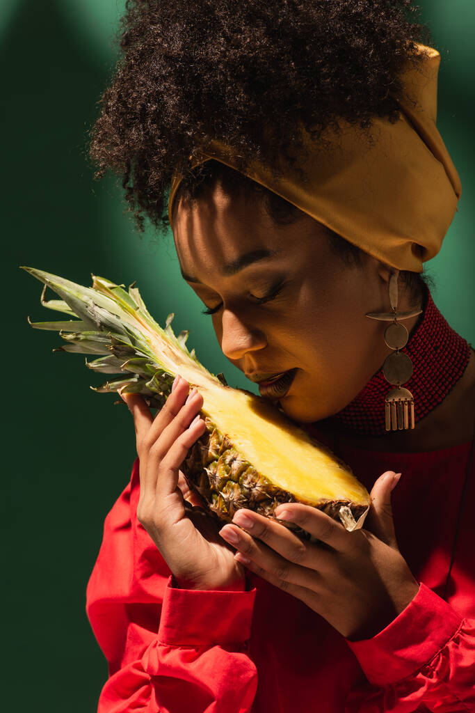νεαρή Αφροαμερικανή με κλειστά μάτια που κρατάει μισό κομμάτι ανανά στα χέρια της πάνω στο πράσινο. - Φωτογραφία, εικόνα